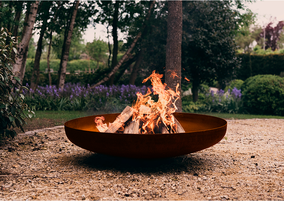 Fogón circular de hierro oxidado con fuego encendido, rodeado de jardín con árboles y flores moradas.